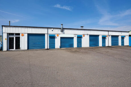 A row of white industrial storage units with closed blue roller doors on a paved lot under a clear blue sky.