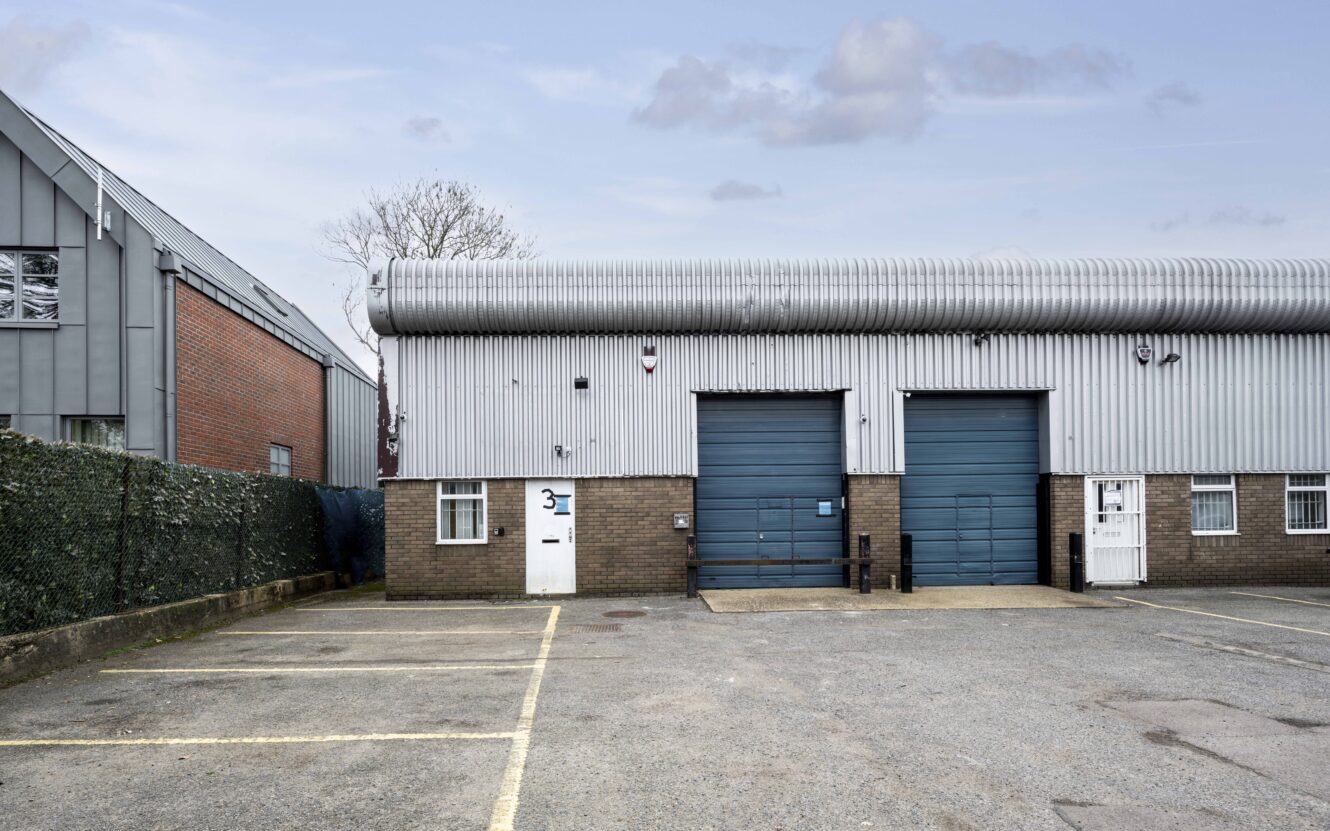 Industrial building with two blue roller shutter doors, a small entrance door, and marked parking spaces in front. The exterior is metal and brick, with a security fence on the left.