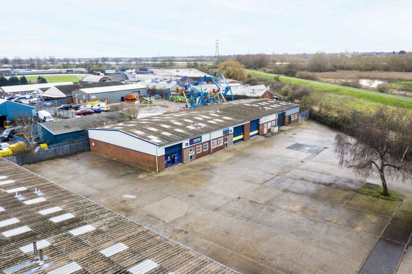 Aerial view of a small industrial complex with several warehouses, a large empty parking lot, and surrounding greenery on a cloudy day.