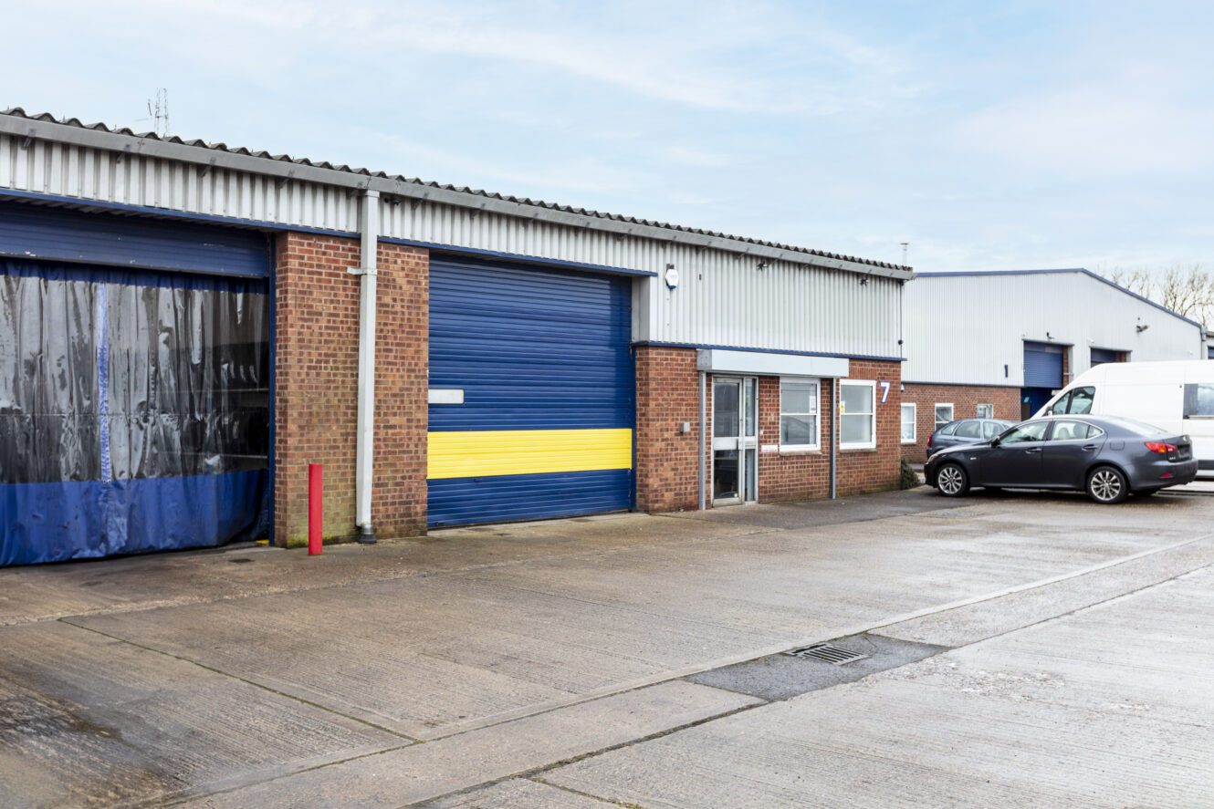 Industrial warehouse with a blue and yellow roller door, brick exterior, glass entrance, parked car, and a white van in front on a concrete lot.