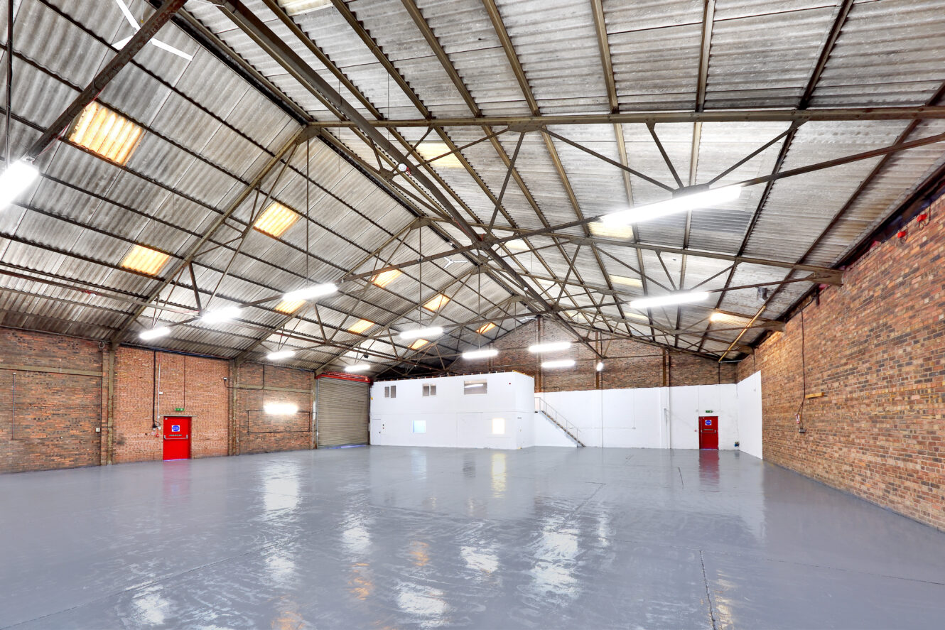 Large empty warehouse with high corrugated metal roof, brick walls, polished gray floor, fluorescent lighting, and small white office structure inside.