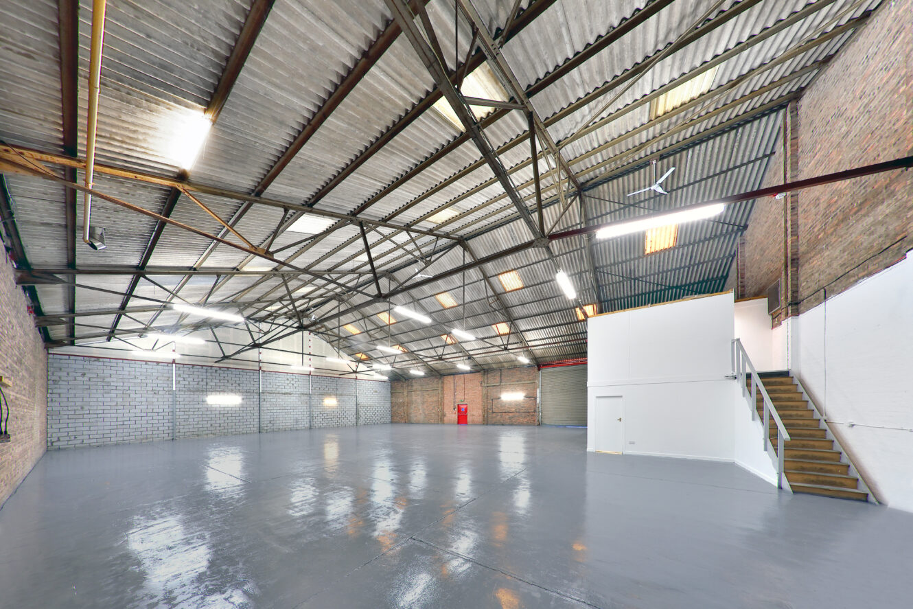 Large empty warehouse with high corrugated metal ceiling, brick walls, polished concrete floor, and a white mezzanine accessed by stairs on the right.
