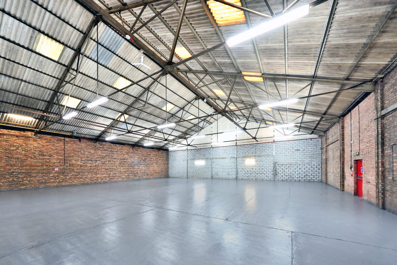 Large empty industrial warehouse with brick and cinder block walls, high ceiling with exposed beams, fluorescent lighting, and a red door.