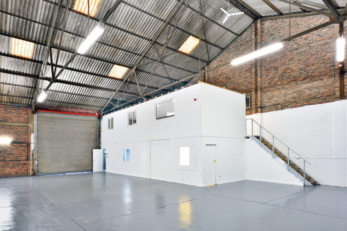 Spacious, empty warehouse interior with high ceilings, exposed brick walls, large roller door, and a white two-story office structure with stairs.