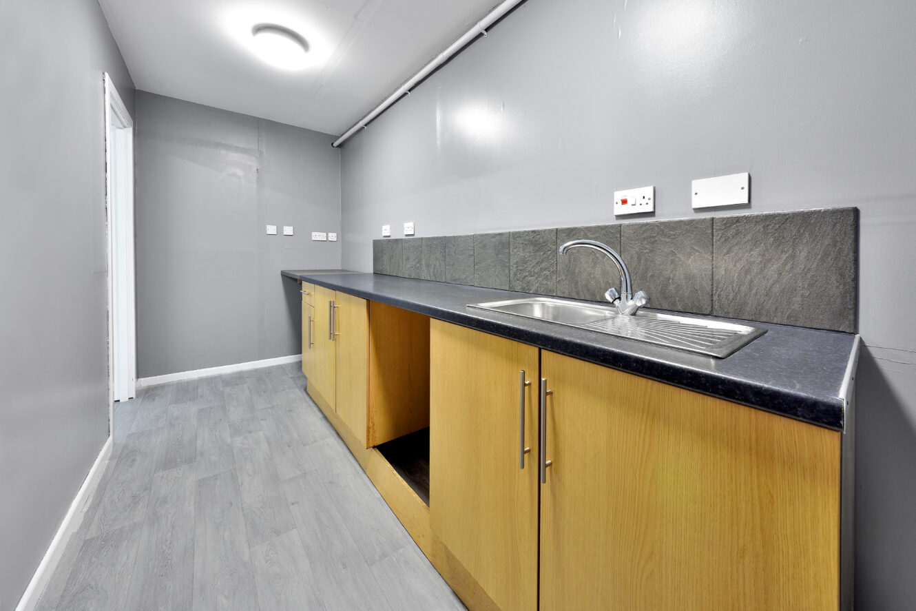 A narrow kitchen with grey walls, wood cabinets, a black countertop, a stainless steel sink, and grey tile backsplash. Some cabinet doors are missing. Ceiling light is on.