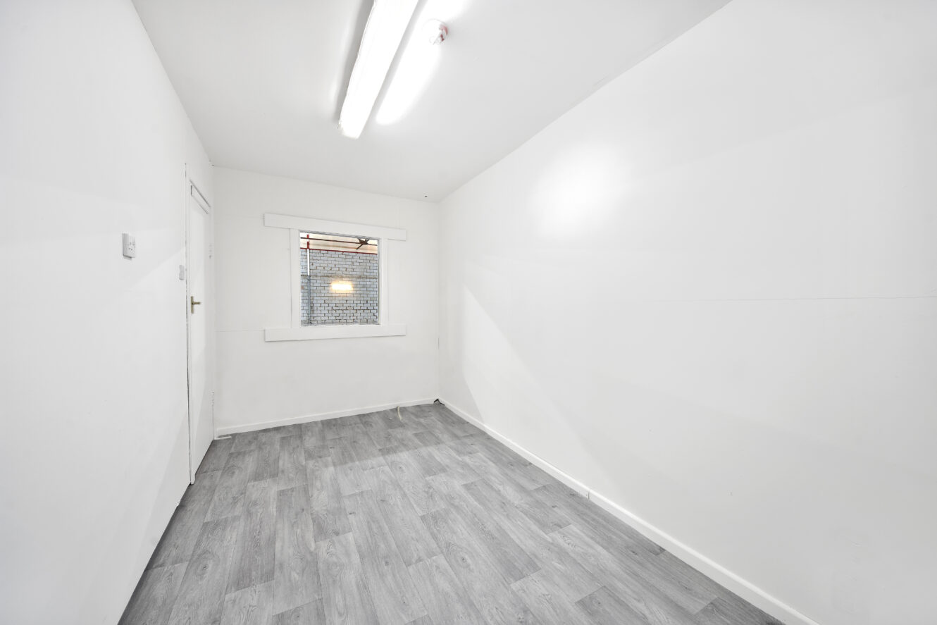 A small, empty room with white walls, gray wood flooring, a closed door, a window, and a fluorescent ceiling light.