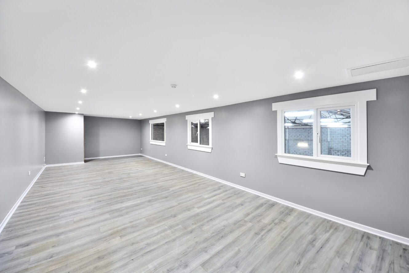 Spacious empty room with gray walls, light wood flooring, recessed ceiling lights, and two windows on the right wall.