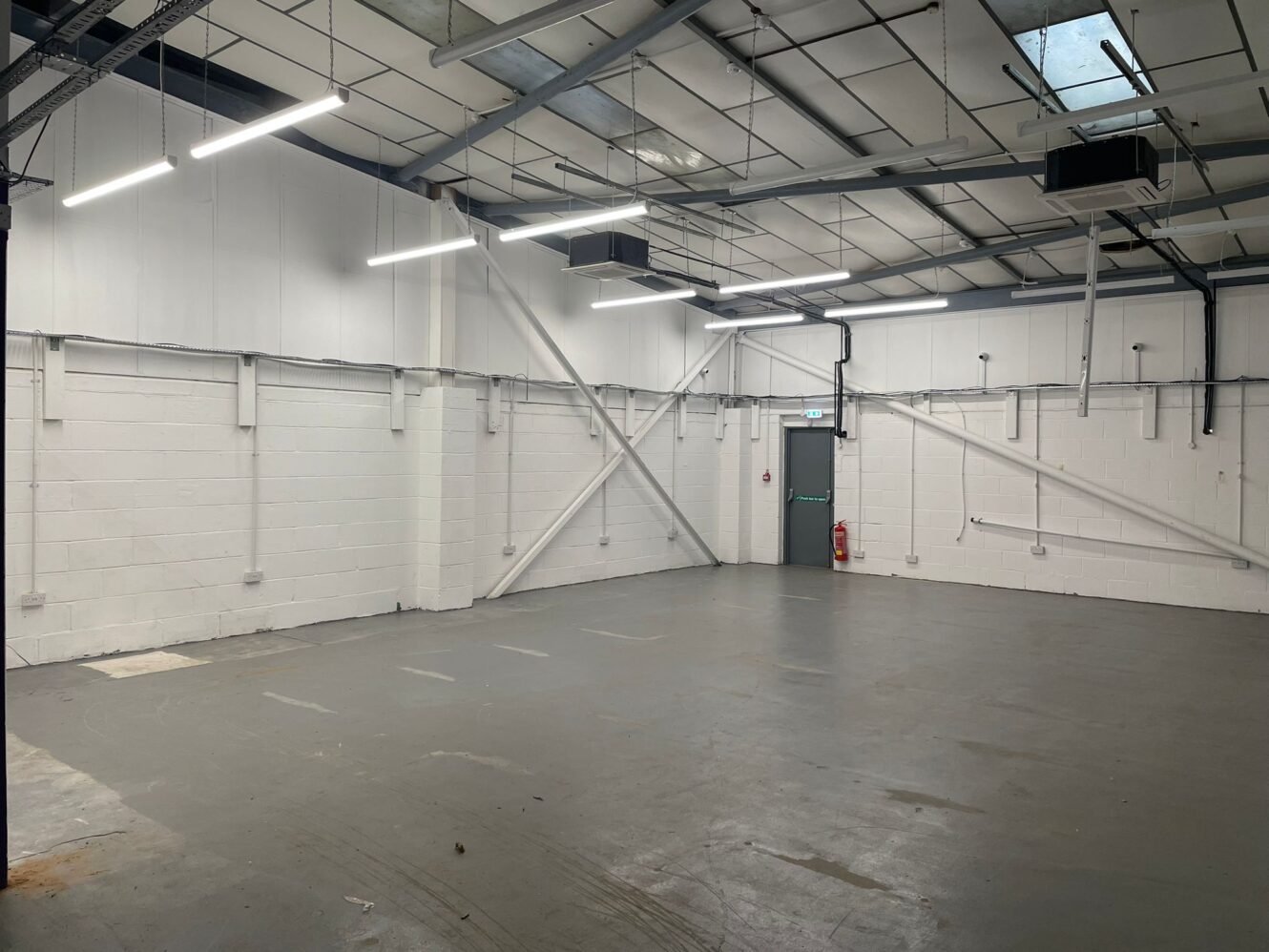 Empty industrial warehouse space with white brick walls, grey concrete floor, exposed ceiling beams, fluorescent lighting, and a metal door at the back.