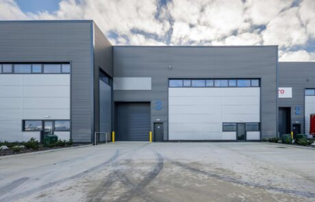 Gray industrial warehouse building with large garage door and smaller entry door, numbered “3,” on a wide, empty concrete lot under a partly cloudy sky.