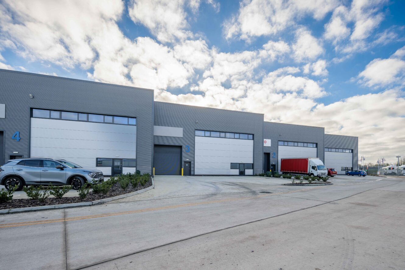 A row of industrial warehouse units with numbered doors, parked cars, and a red cargo truck under a partly cloudy sky.
