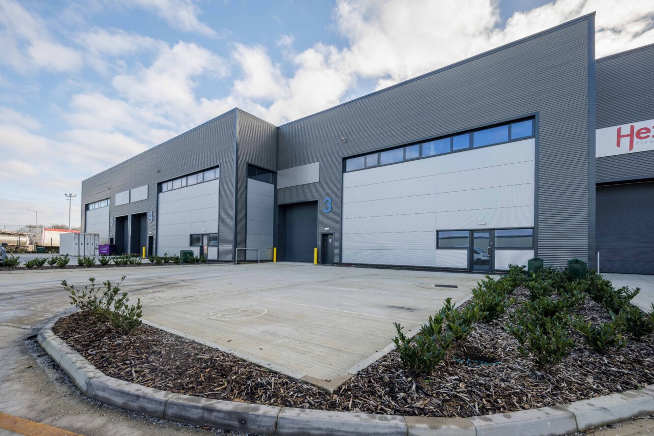 A modern industrial warehouse with large grey doors, numbered 3, and minimal landscaping in front, under a partly cloudy sky.