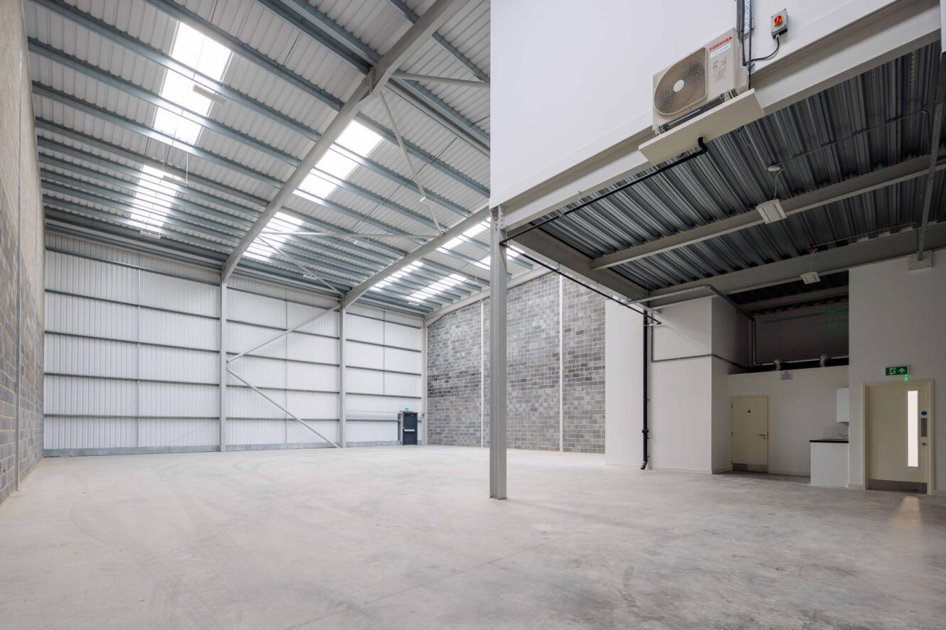 Large, empty industrial warehouse with high metal ceiling, exposed beams, concrete floor, and a small office area in one corner.