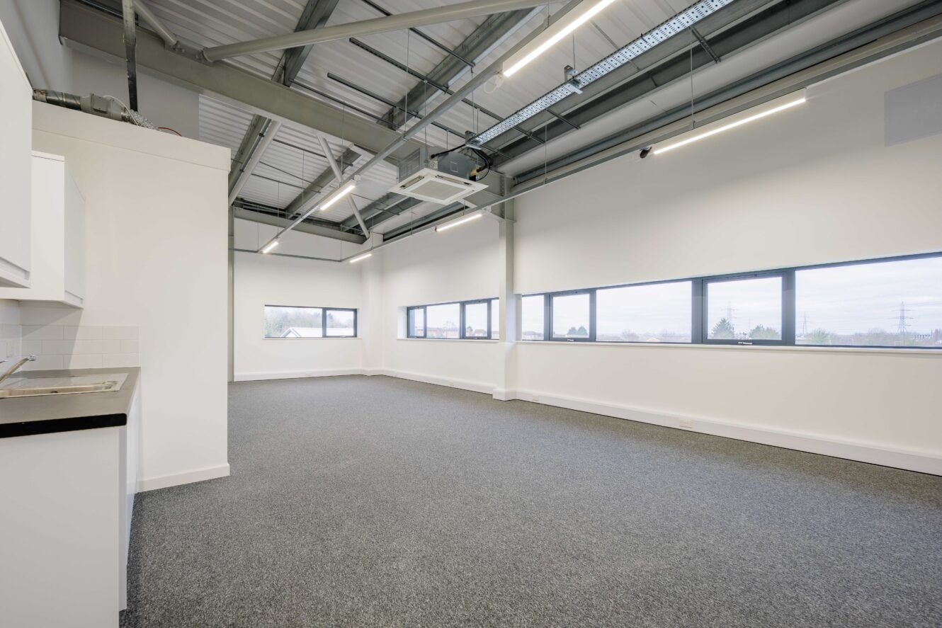 Empty, modern office space with gray carpet, white walls, exposed ceiling beams, fluorescent lighting, and large windows along one side letting in natural light.