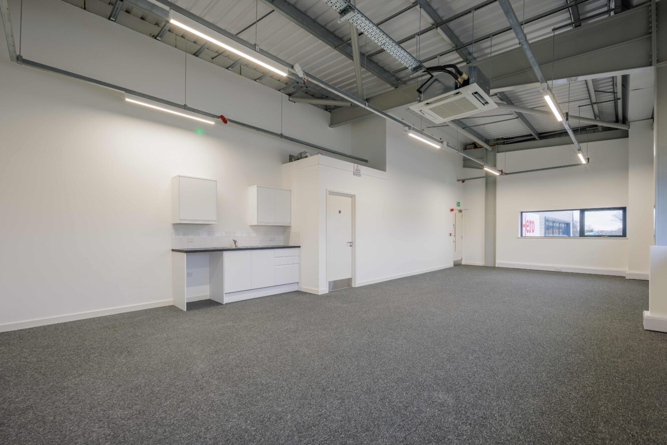 An empty modern office space with grey carpet, white walls, exposed ceiling, a kitchenette, and large windows allowing natural light.