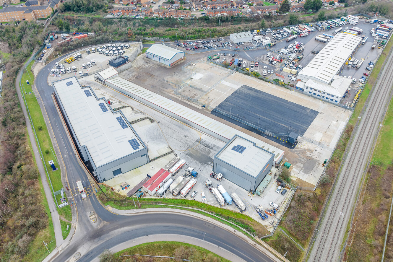 Aerial view of an industrial site with warehouses, trucks, parked vehicles, and adjacent railway tracks, surrounded by roads and greenery.