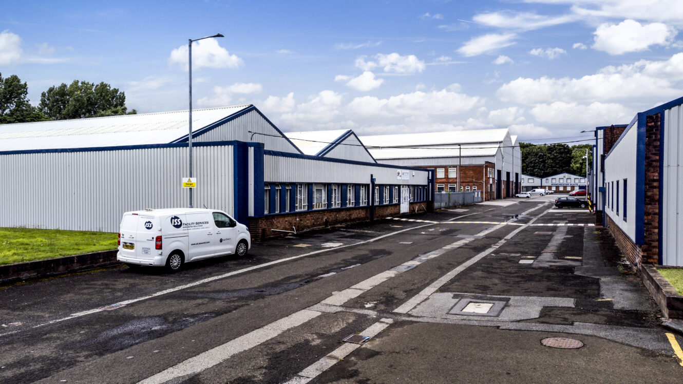 A white van is parked beside a large industrial warehouse complex on an empty street under a partly cloudy sky.