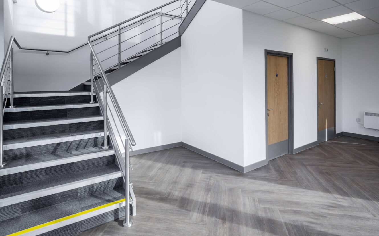 A modern indoor stairwell with gray steps, metal railings, two closed wooden doors, and a caution tape marking at the base of the stairs.