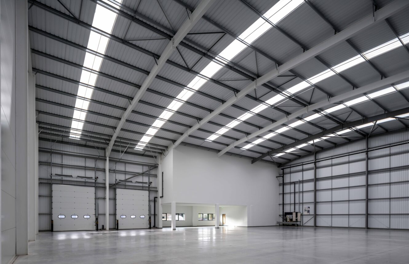 Spacious empty warehouse with high ceilings, skylights, large industrial doors, and a small office area built into one corner.