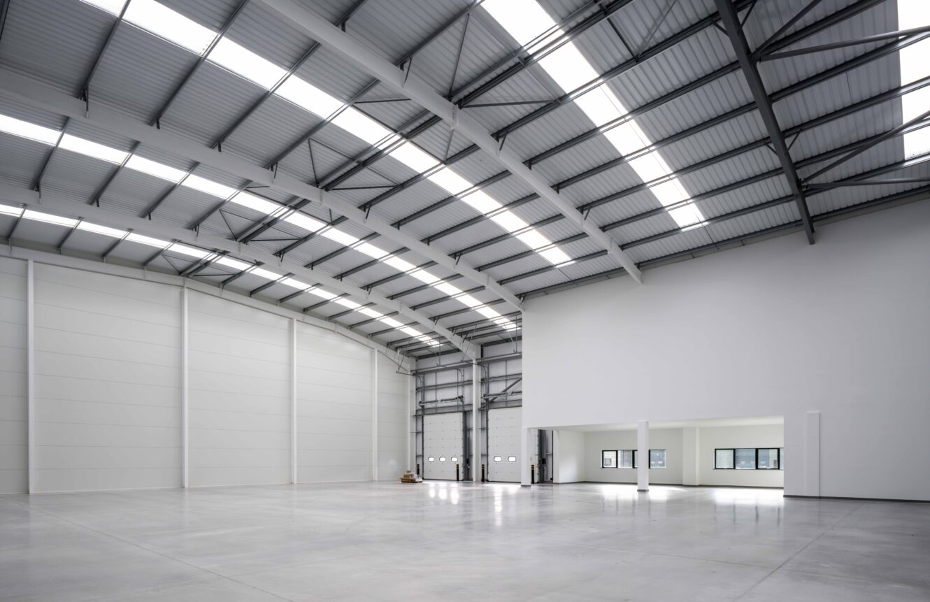 Interior of a large, empty warehouse with high ceilings, exposed beams, polished concrete floor, white walls, and large windows on an elevated office section.