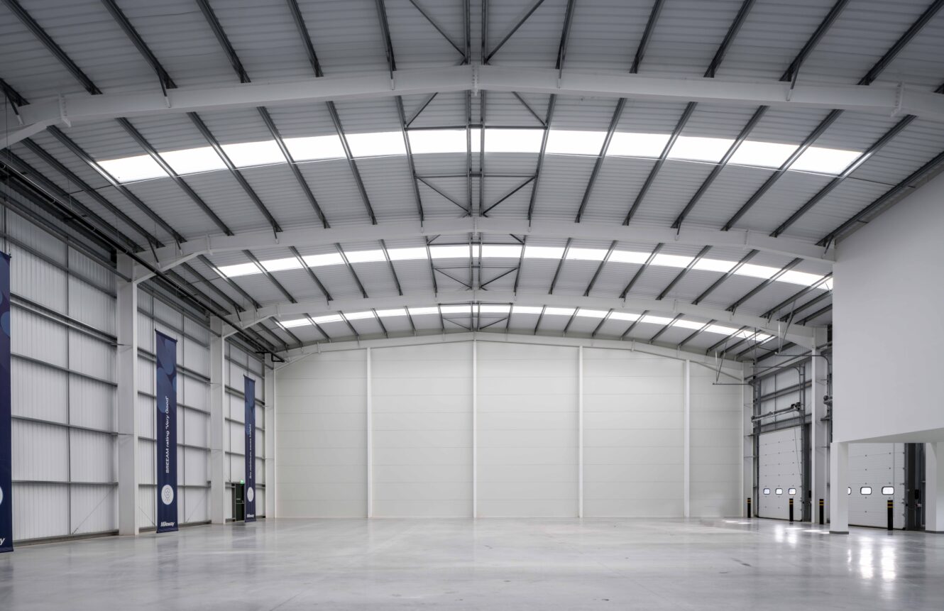Interior of a large, empty warehouse with high ceilings, skylights, white walls, and industrial doors on the right side.