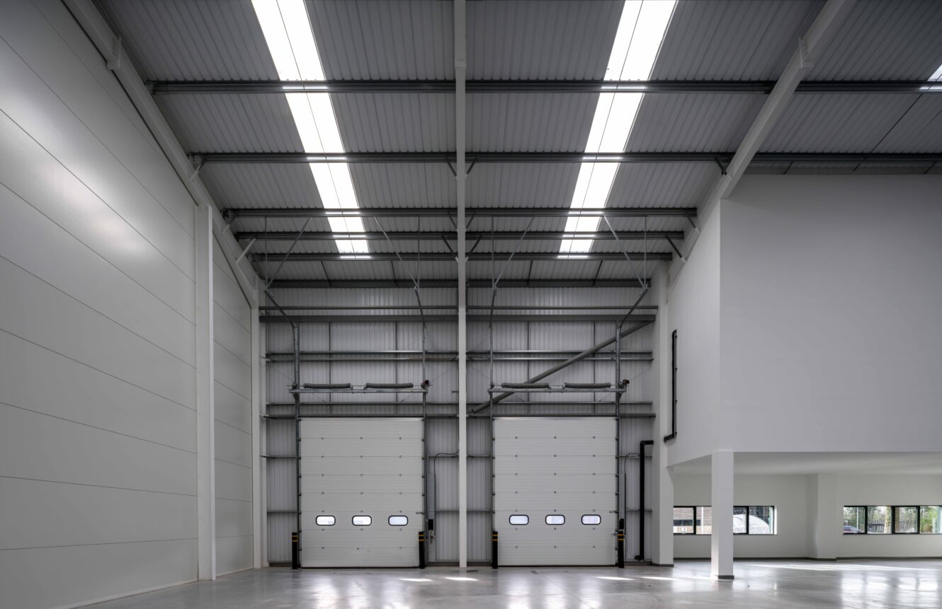 Interior of an empty industrial warehouse with high ceilings, two large loading bay doors, and a sectioned office area with windows on the right.