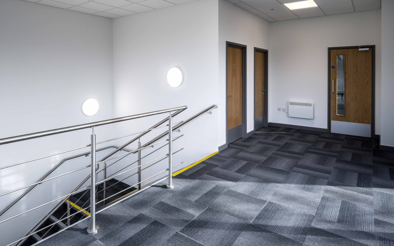A modern indoor staircase with metal railings and grey carpeted floors, leading to a hallway with multiple wooden doors and circular windows on the wall.