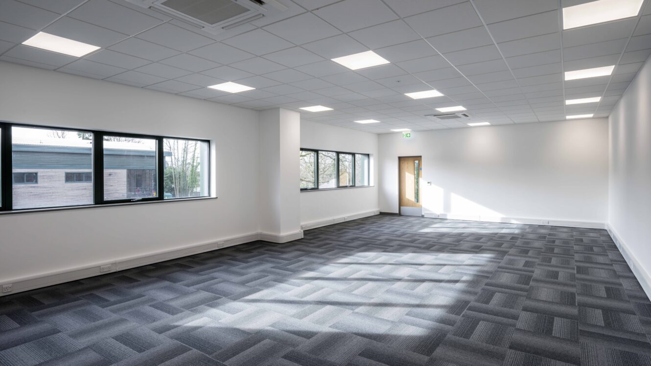 An empty office room with patterned carpet, white walls, large windows, and ceiling lights. Sunlight enters through the windows near a wooden door.