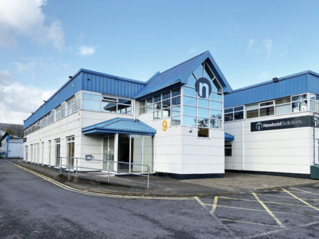 A modern two-story office building with blue roofing, large windows, and the sign Newbold Solicitors on the facade. A ramp and yellow number 9 are visible near the entrance.