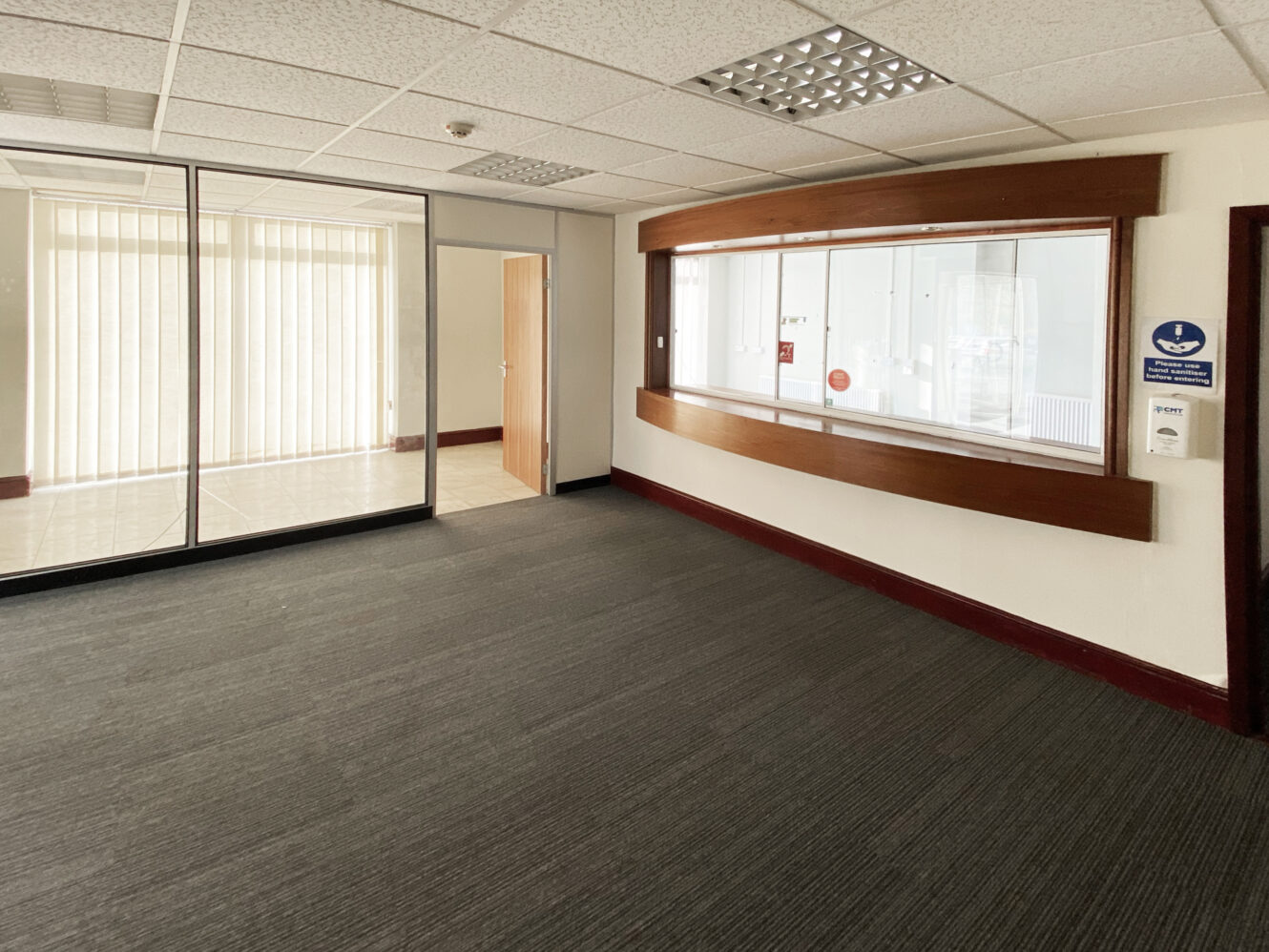 Empty office space with gray carpet, glass partition, open wooden door, large windowed counter, and fluorescent ceiling lights.
