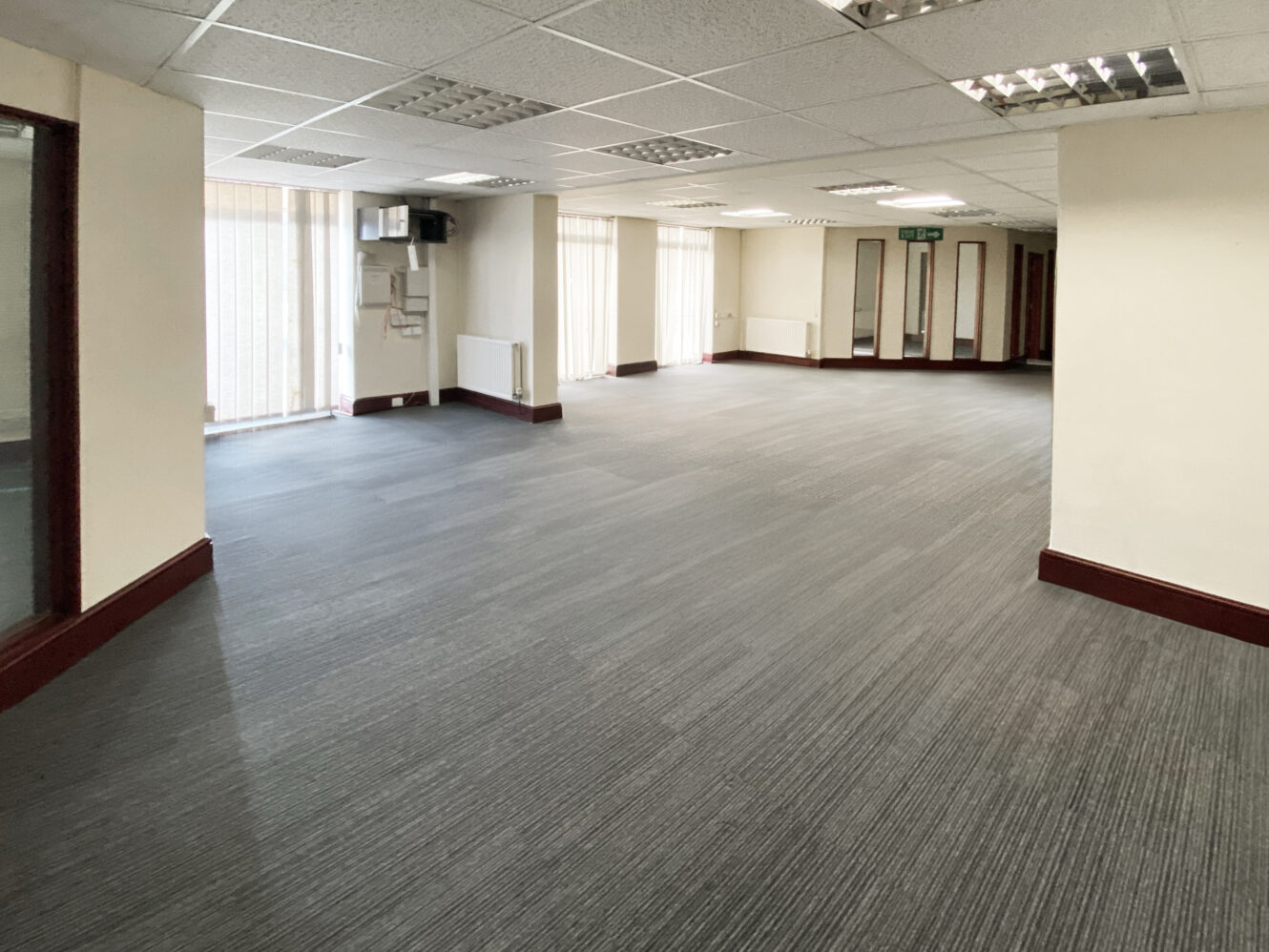 Large, empty office space with gray carpet, cream walls, glass doors, and fluorescent ceiling lights. Multiple windows allow natural light into the room.