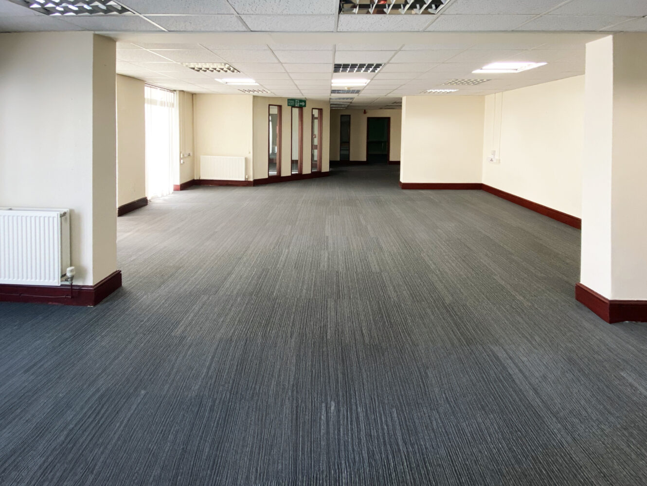 Spacious, empty office room with grey carpet, cream-colored walls, and fluorescent ceiling lights.