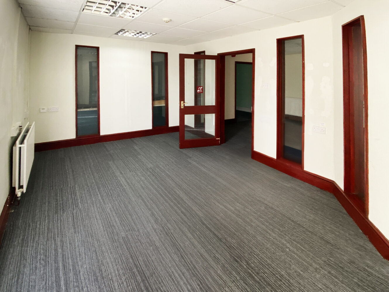 Empty office room with gray carpet, white walls, maroon trim, glass windows, ceiling tiles, and an open glass door.