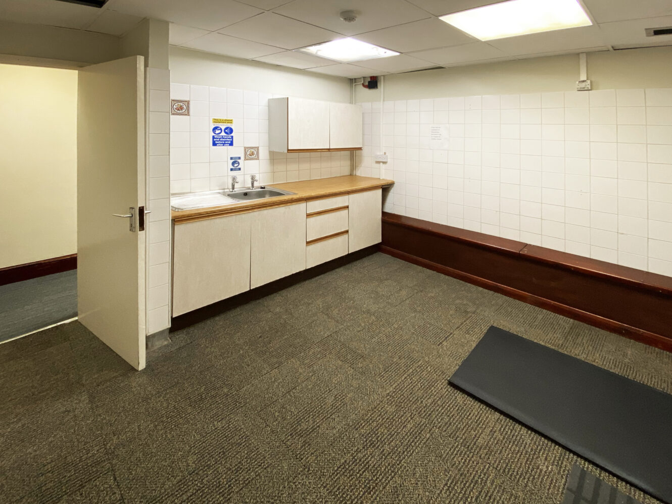 A small room with a kitchenette, white cabinets, a sink, tiled walls, open door, and a black mat on the carpeted floor.