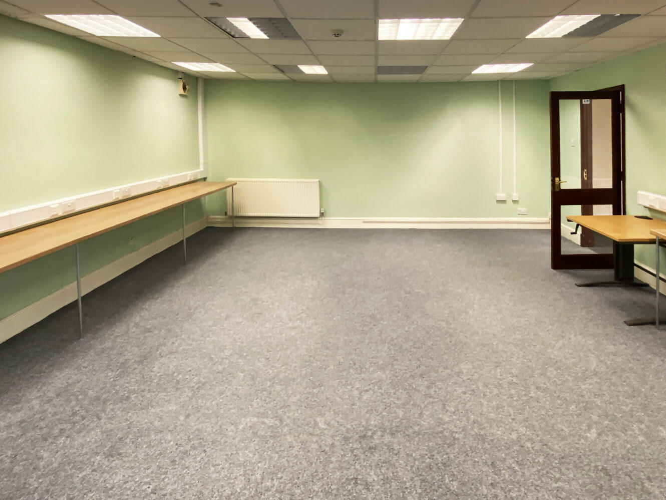 Empty office room with light green walls, grey carpet, fluorescent ceiling lights, long wooden desks along the sides, and an open door to the right.