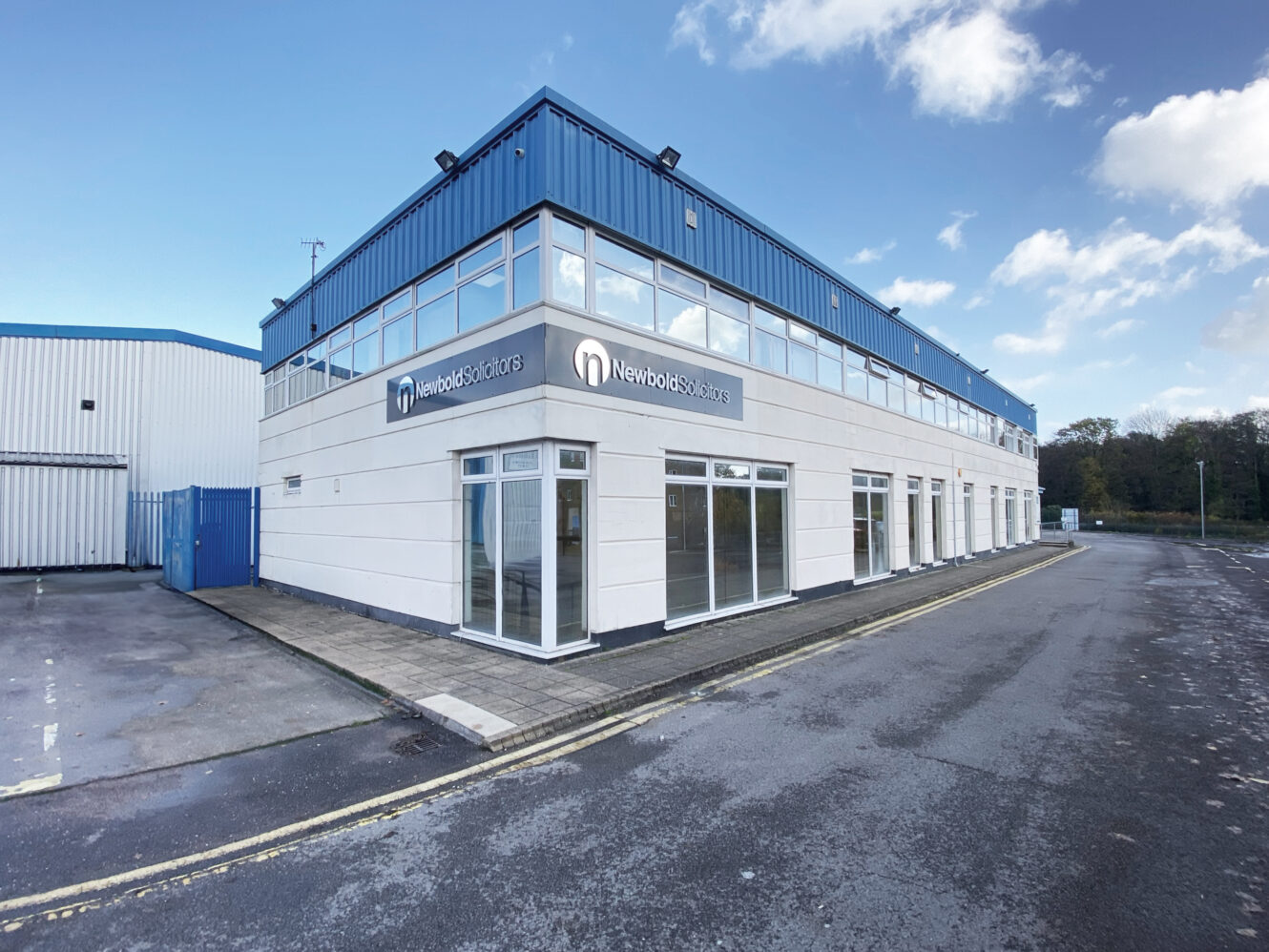 A modern two-story commercial building with Newfold Solutions signage, large windows, and a blue roof, located by an empty road on a clear day.