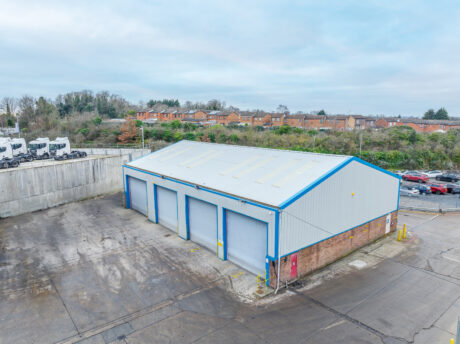Large industrial warehouse with four closed roller doors, surrounded by a paved lot, with houses and greenery visible in the background.