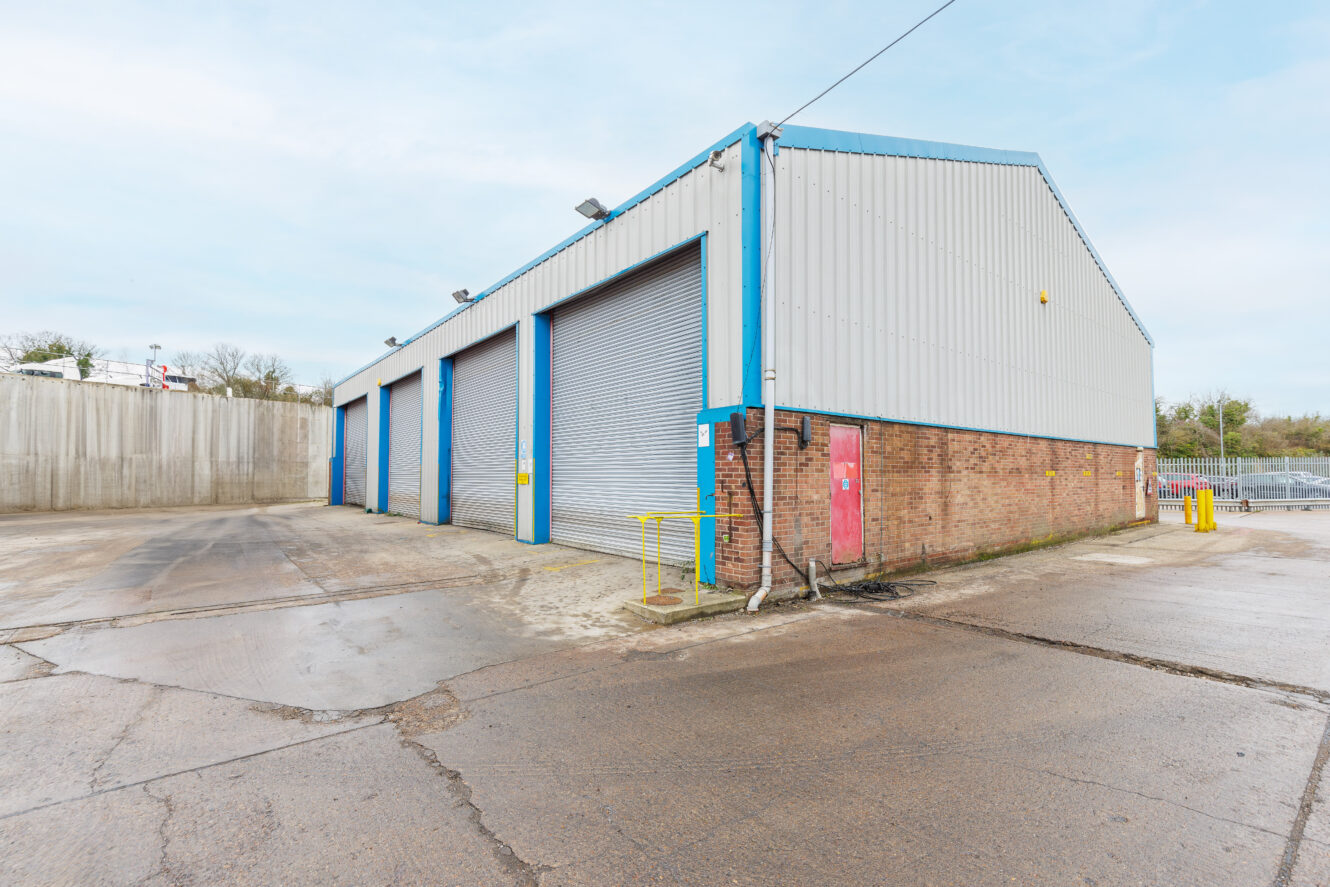 A large industrial warehouse with three gray roller shutter doors, brick and metal siding, and blue trim, situated on a concrete lot.