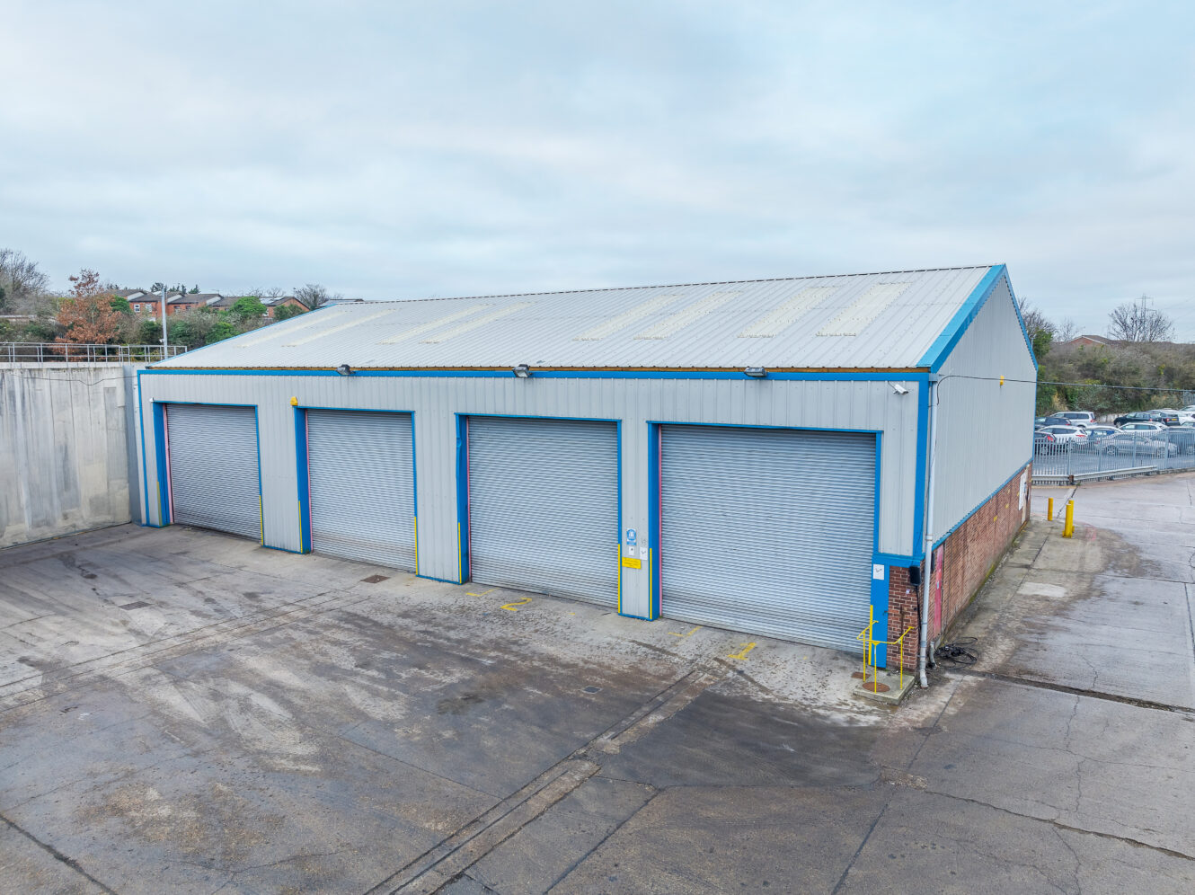 A large industrial building with four closed roller shutter doors, surrounded by a concrete yard and parking area on a cloudy day.