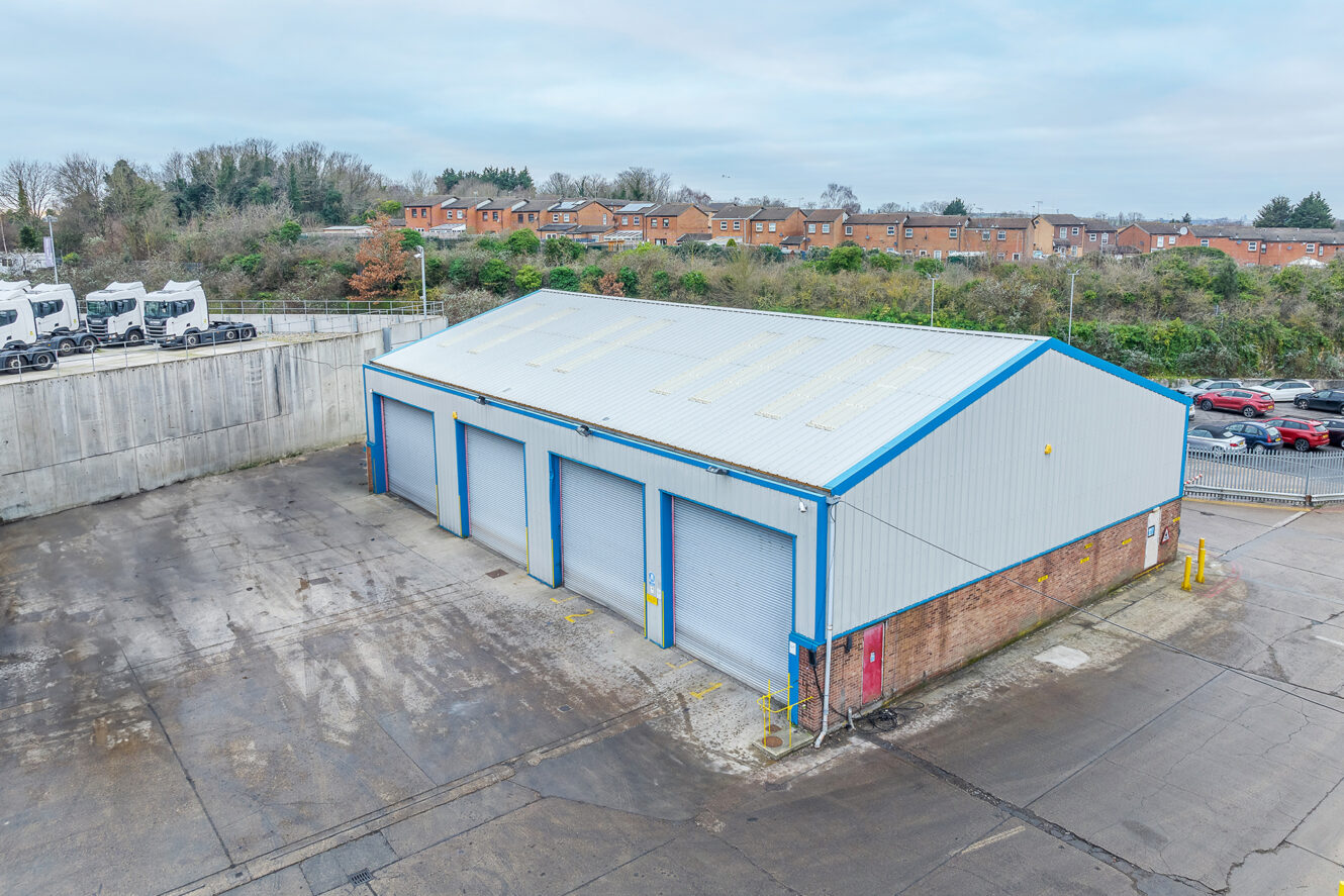 A large industrial warehouse with four blue roller doors, surrounded by a paved lot, cars, and trees, with houses visible in the background.