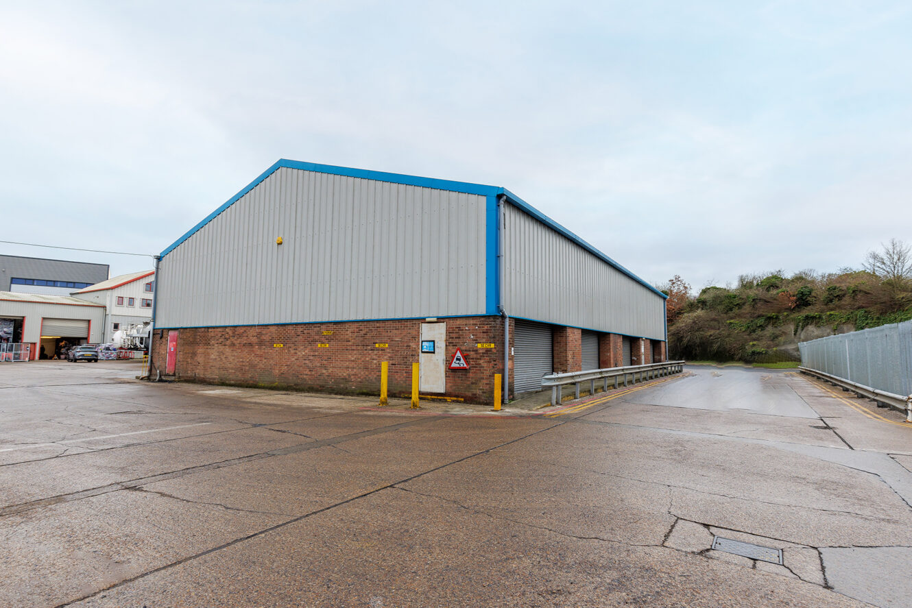 A large industrial warehouse with brick and metal siding, surrounded by a paved lot and safety barriers, under a cloudy sky.