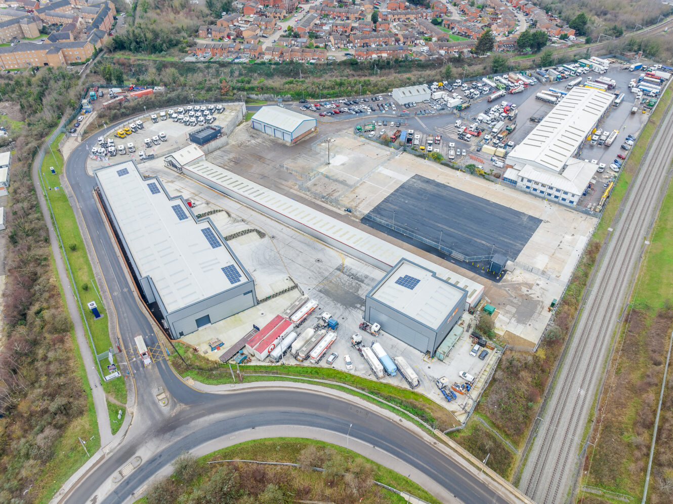Aerial view of an industrial complex with multiple warehouses, parking lots, delivery trucks, and adjacent railway tracks, bordered by a residential area and curved roads.