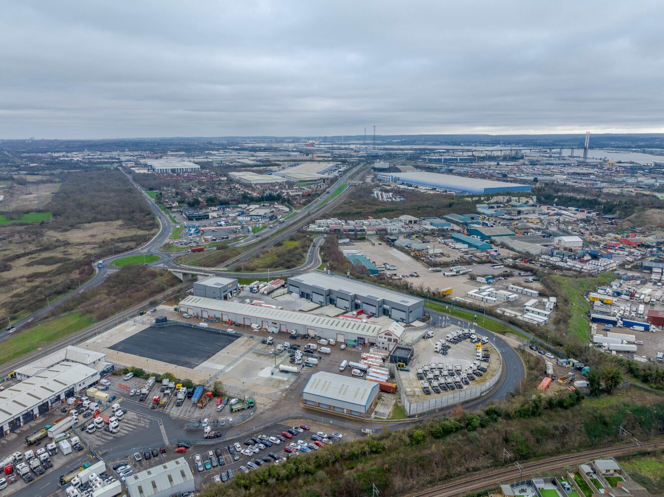 Aerial view of an industrial estate with warehouses, trucks, and assorted vehicles under an overcast sky, surrounded by roads and patches of greenery.