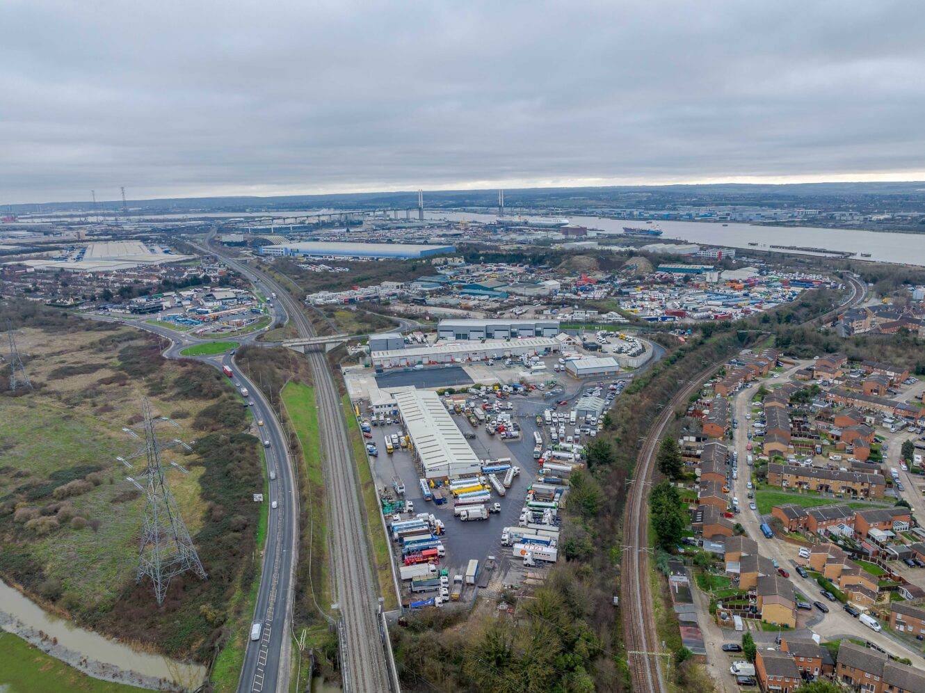 Aerial view of an industrial area with warehouses, parked trucks, roads, nearby houses, and a river in the background under a cloudy sky.