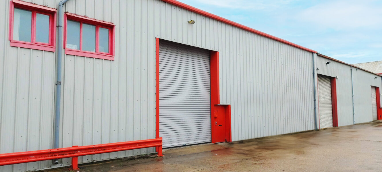 A large industrial building with grey corrugated metal walls, red trim, and multiple roller shutter doors on a wet concrete surface.