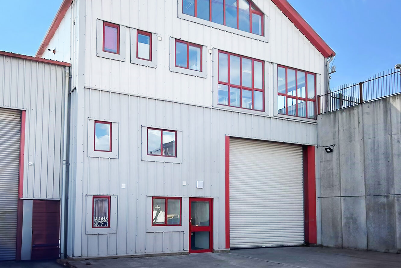 A modern industrial building with red-trimmed windows, a large roller shutter door, a concrete yard, and a high concrete wall on the right.
