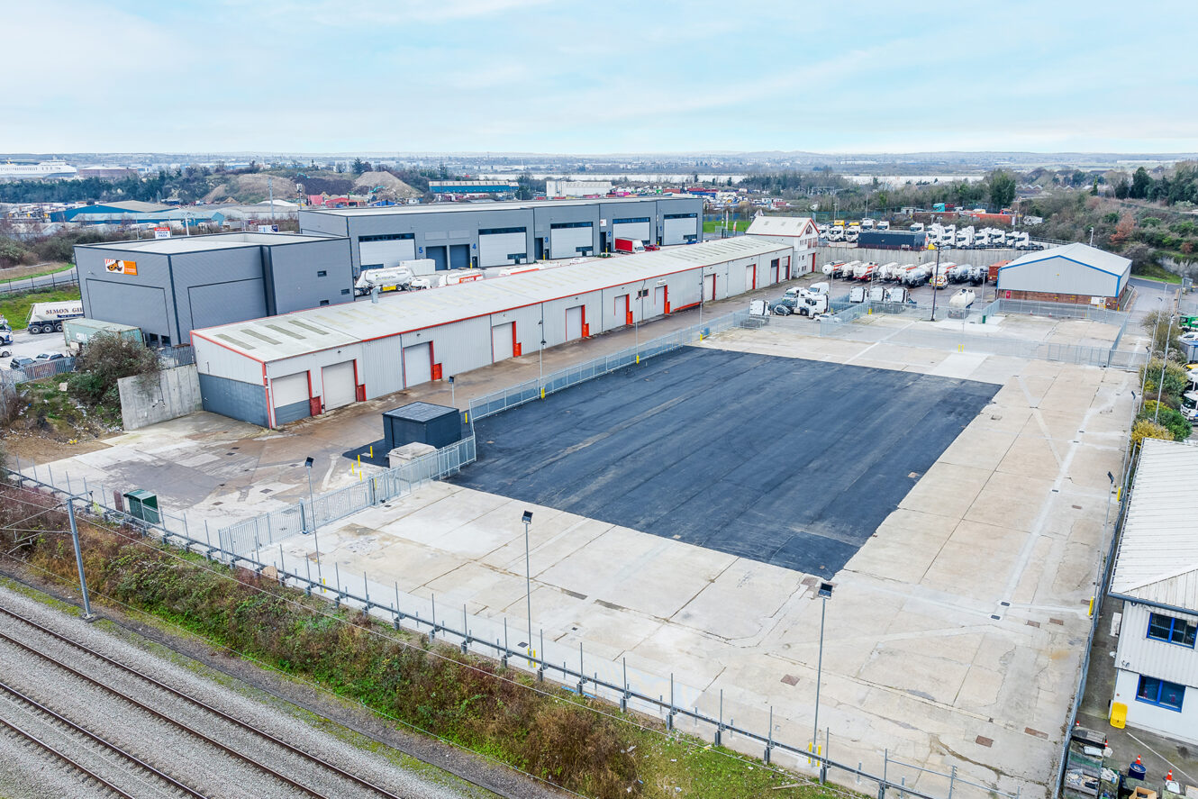 Aerial view of an industrial warehouse complex with multiple loading bays, a fenced paved yard, parked trucks, and adjacent rail tracks.