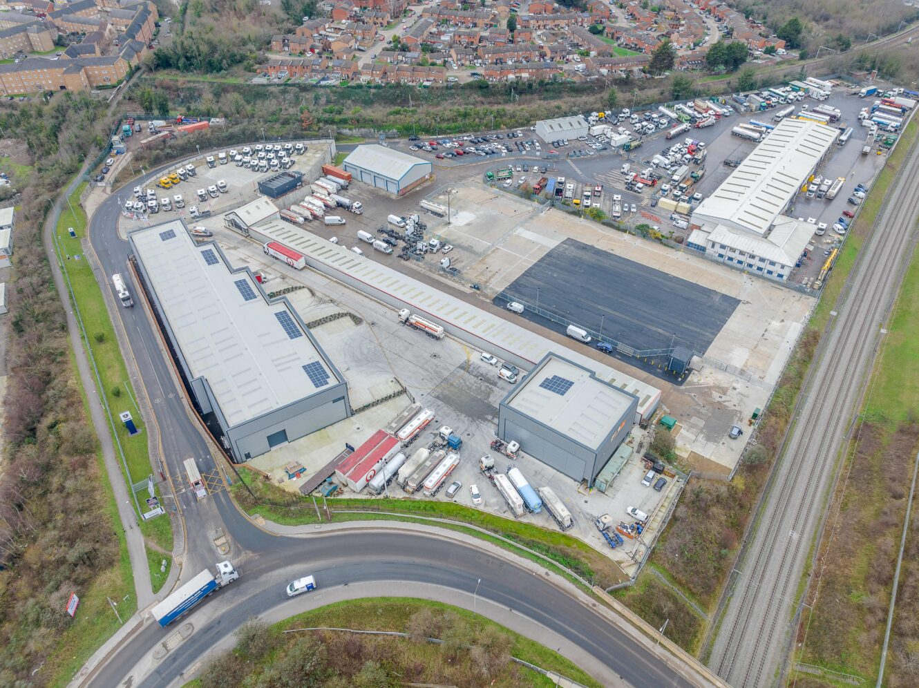 Aerial view of an industrial complex with several large warehouses, parked trucks, and nearby roads; residential area visible in the background.