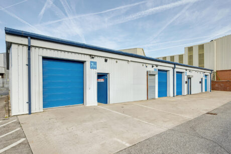 A row of industrial units with blue roller doors and white walls, each with numbered signs, set beside a concrete parking area under a clear sky.