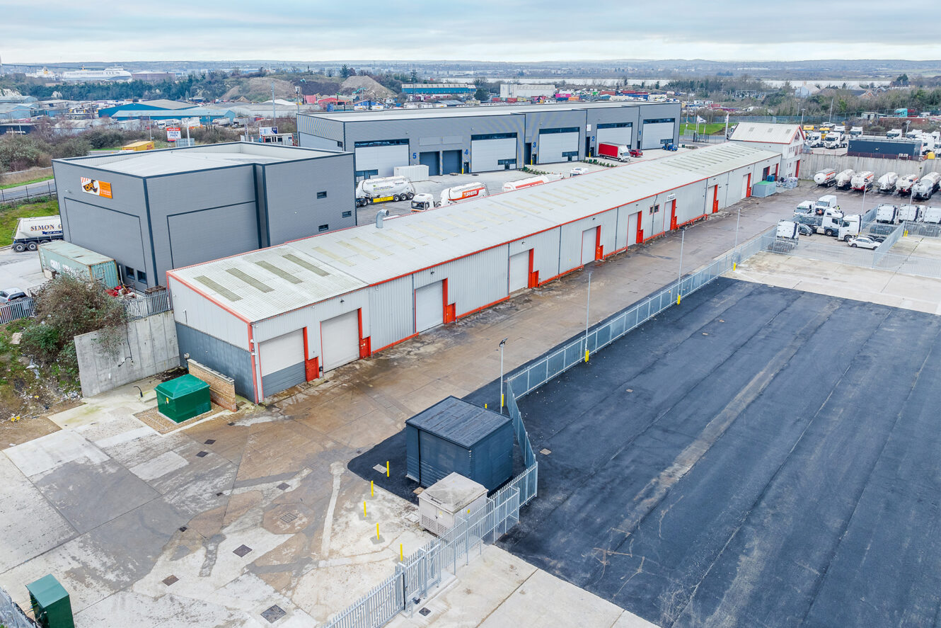 Aerial view of an industrial site with warehouse buildings, several trucks, parked vehicles, and a fenced area under cloudy sky.