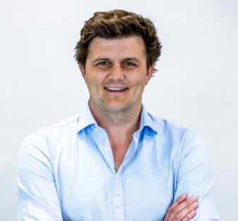 A man with brown hair in a light blue button-up shirt stands in front of a plain white background, smiling with his arms folded.